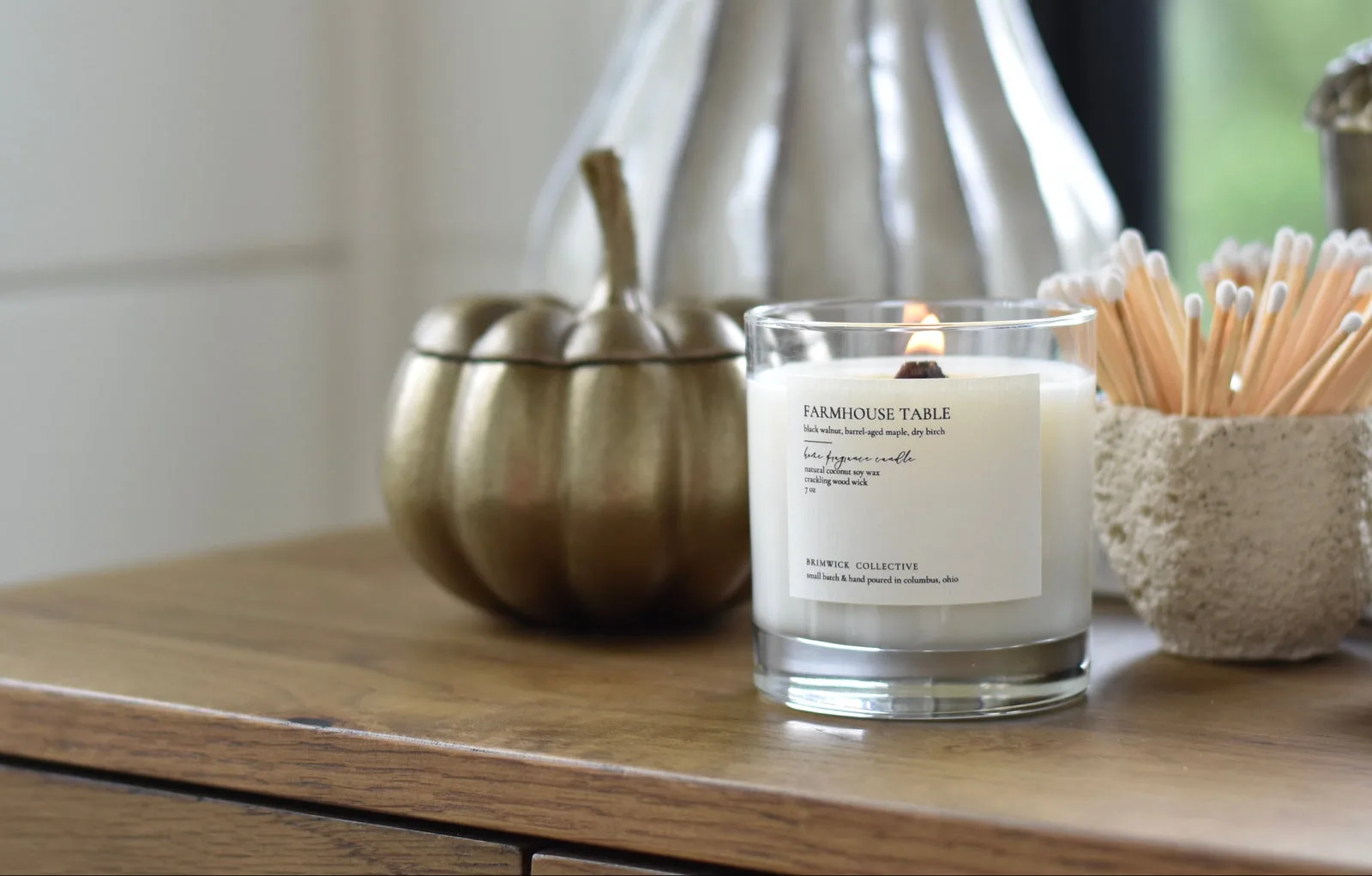A Brimwick Collective Farmhouse Table candle with a wood wick burning beside a brass pumpkin
