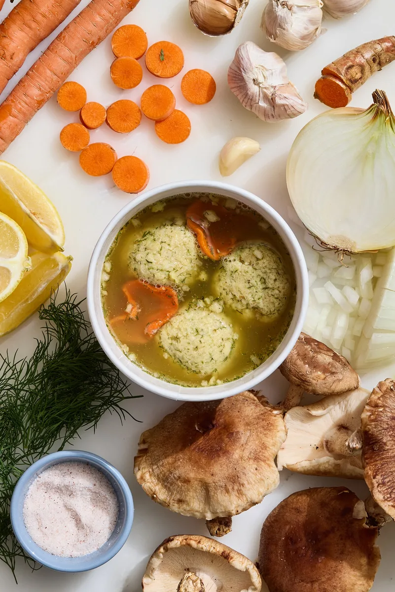 A bowl of matzo ball soup with carrots, dill and the ingredients around it