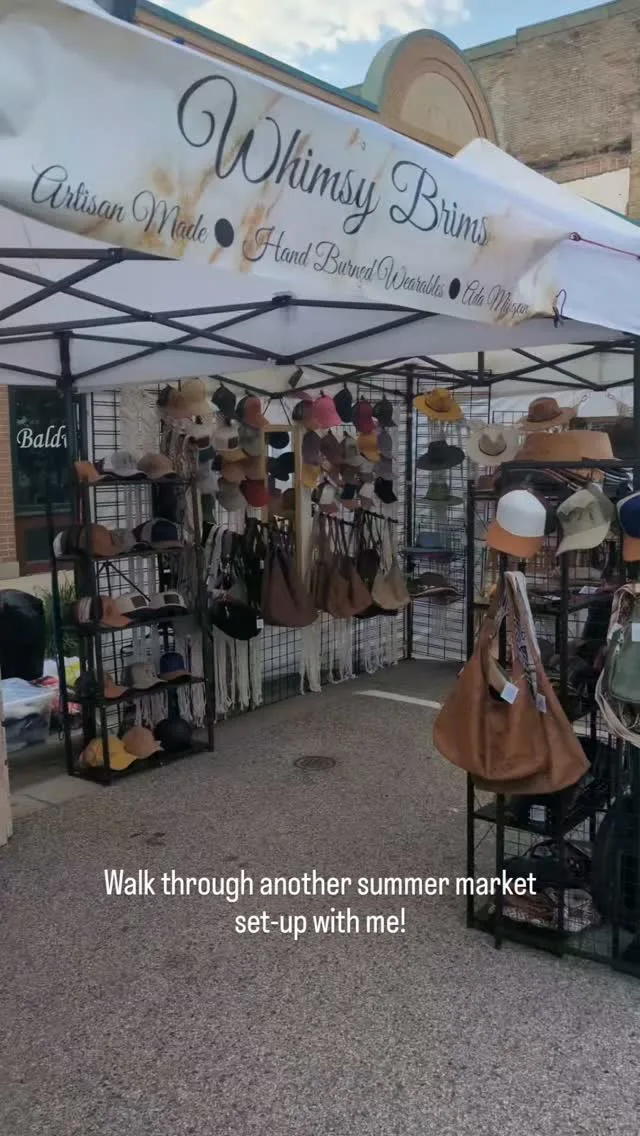The Whimsy Brims booth at a summer market, walls of hand-burned hats and purses